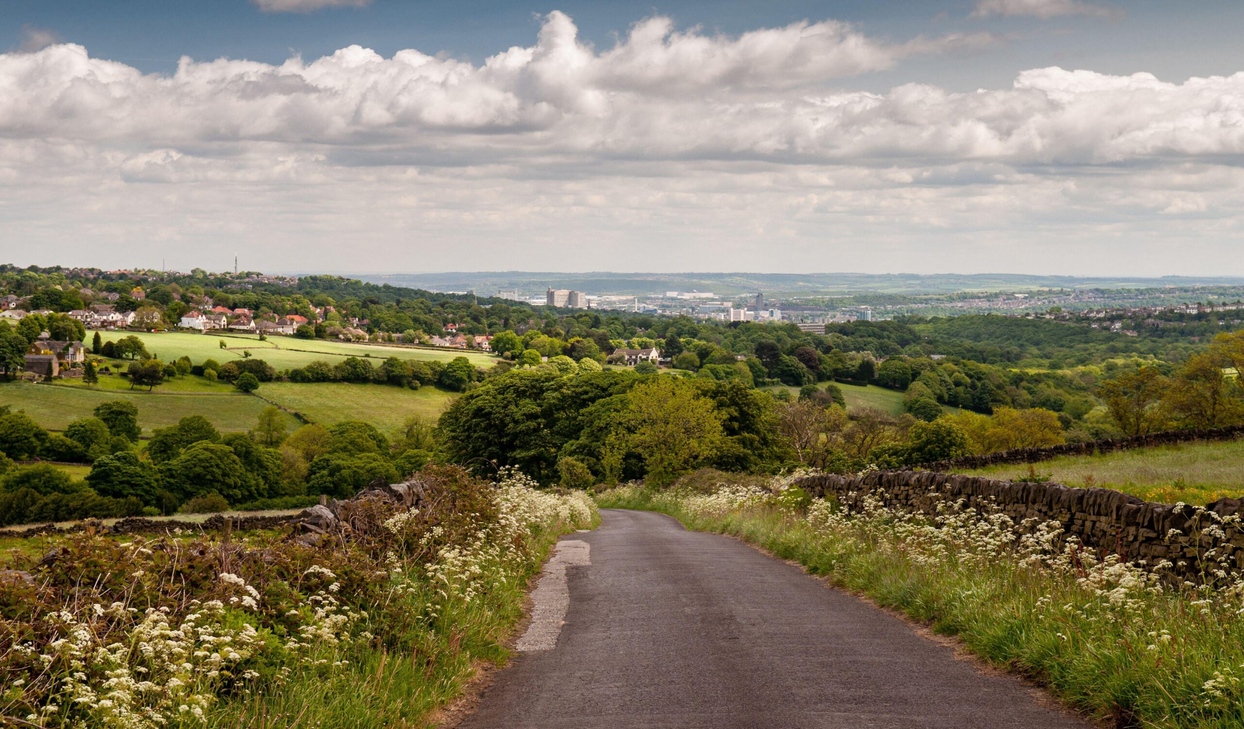 A country lane descends from the Peak District hills into the Mayfield Valley and the suburbs of Sheffield, with the city centre cityscape in the dist