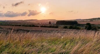 A sun setting over the East Yorkshire countryside, with light fluffy clouds in the sky, rolling hills in the background and a meadow in the foreground.